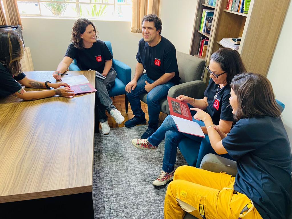 Fotografia colorida de uma reunião com o time da CASA consultoria. Em semicírculo e sentados em poltronas, da esquerda para direita, Mariane, Natalia, Allan, Andréia e Juliana. todos vestem as camisetas da empresa. Estão em uma sala iluminada. À esquerda, uma mesa de madeira. Ao centro, na parte inferior da imagem, um tapete cinza. No canto superior direito, uma estante de livros e ao fundo, parede branca e a parte inferior de uma janela de vidro.