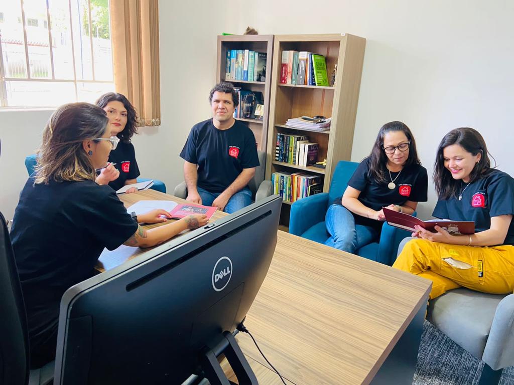 Fotografia colorida de uma reunião com o time da CASA consultoria. Em semicírculo, sentados em poltronas, da esquerda para direita, Mariane, Natalia, Allan, Andréia e Juliana. Todos usam as camisetas pretas com a logomarca da empresa do lado esquerdo do peito. Ao centro e entre os integrantes, uma mesa de madeira. Sobre a mesa, no canto inferior esquerdo da imagem, um computador preto. No canto superior esquerdo, uma parte de uma janela de vidro. Ao fundo e centralizado, uma estante de livros.