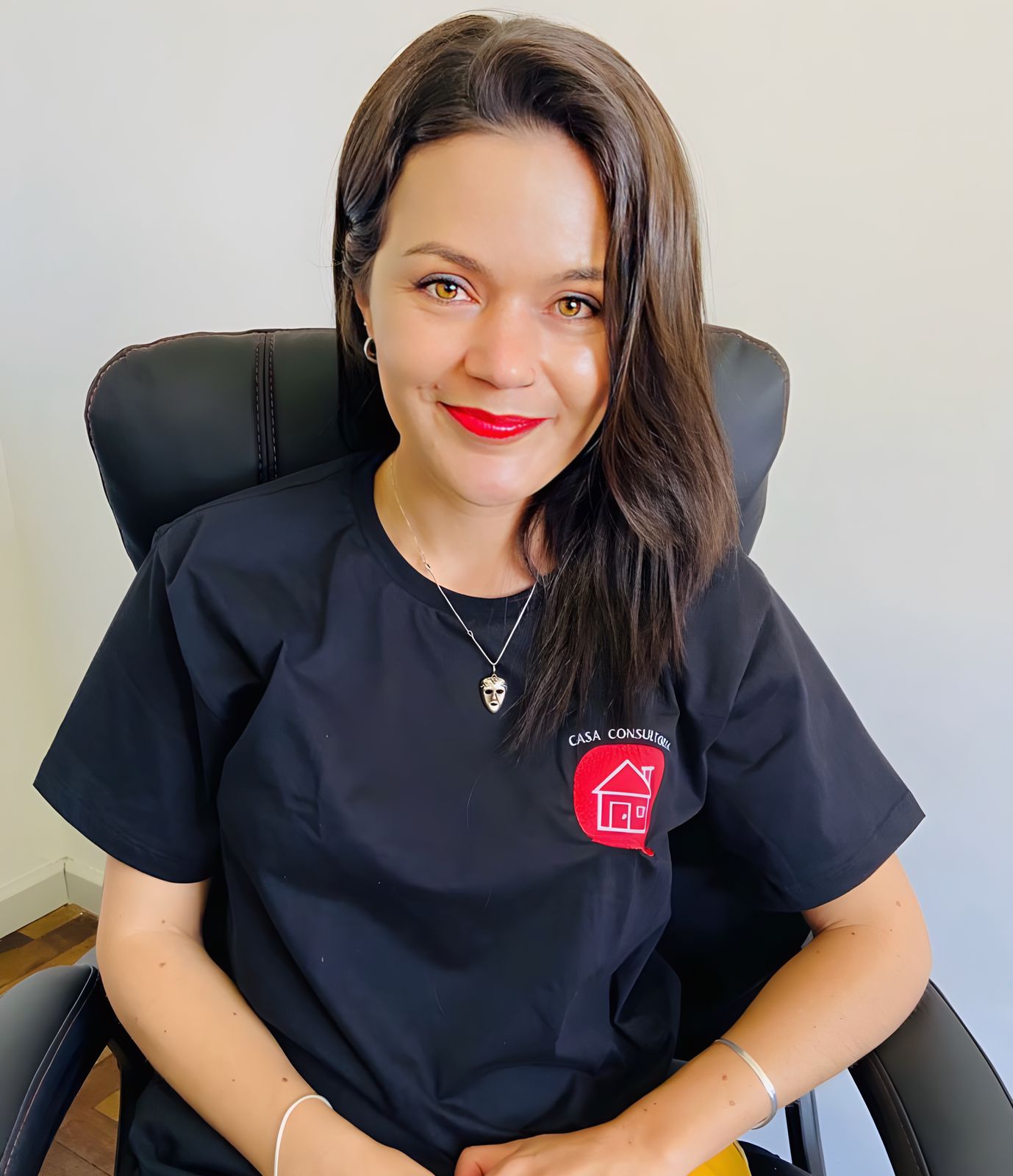 Fotografia colorida de Juliana Partyka. Mulher com pele branca, olhos verdes, cabelos castanhos escuros, lisos e jogados ao lado esquerdo do ombro. Usa batom vermelho e sorri. Veste camiseta preta com a logomarca da CASA consultoria do lado esquerdo do peito. Está sentada em cadeira preta com as mãos sobre o colo e os cotovelos apoiados nos braços da cadeira. Utiliza pulseiras nos dois pulsos, colar e brincos na cor prata. Ao fundo, parede branca.