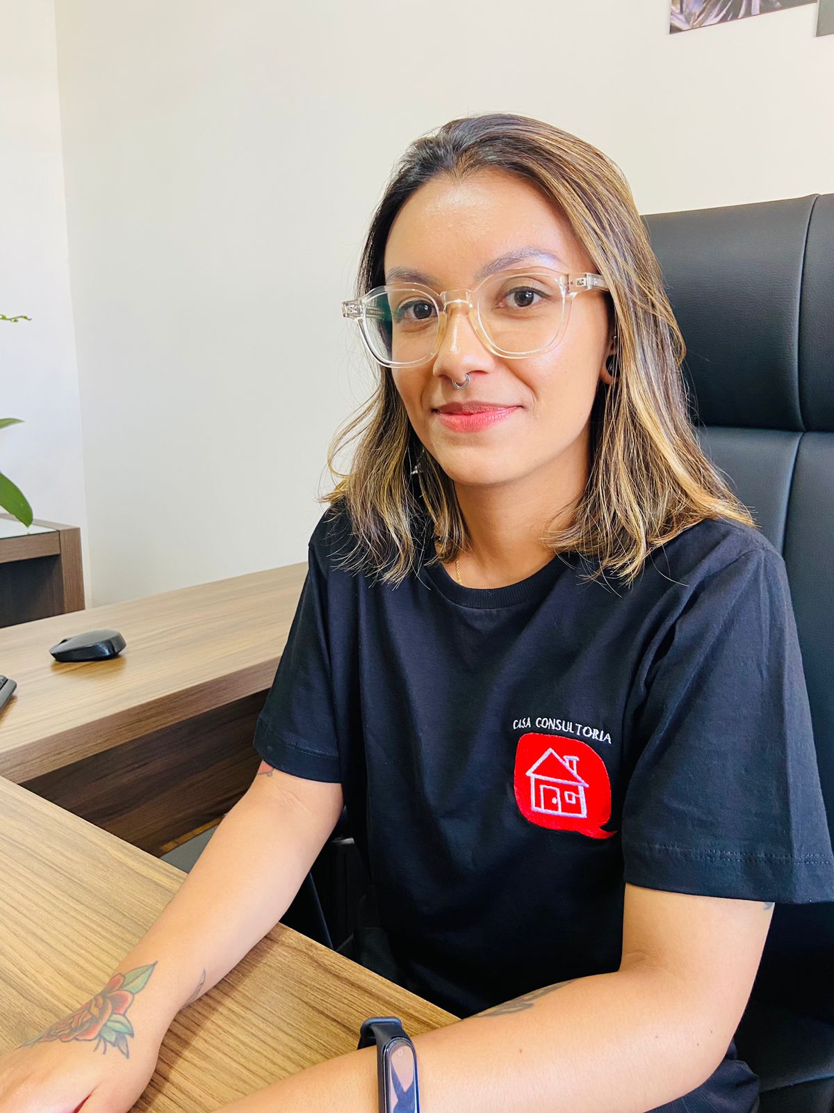 Fotografia colorida de Mariane Laurentino. Mulher de pele parda, cabelos castanhos com mechas claras, curtos e lisos. Veste camiseta preta com a logomarca da CASA consultoria no canto esquerdo do peito, óculos com armação transparente e relógio no pulso esquerdo. Está sentada em uma cadeira preta e sorri. Seus braços estão apoiados em uma mesa de madeira à frente. Do lado esquerdo da imagem, um mouse preto e ao fundo, parede branca.