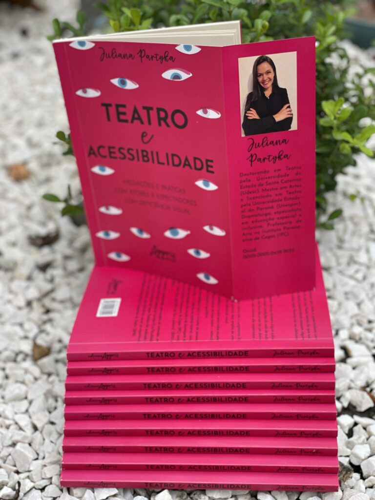 Na imagem, pilha de livros, com a brochura voltada para fora, que acompanha o seguinte texto: Editora Appris, Teatro e Acessibilidade, Juliana Partyka. Em cima do conjunto, livro apoiado com destaque para a capa na cor rosa pink e diversos olhos com púpila azul, espalhados e aba interior voltada para fora, com foto de Juliana Partyka. Juliana tem pele branca, cabelos escuros, na altura dos ombros, usa um casaco preto, está com os braços cruzados e sorri. A capa do livro, apresenta em letras pretas, o texto: Juliana Partyka, Teatro e Acessibilidade: Mediações e Práticas com atores e espectadores com deficiência visual, Appris Editora. 